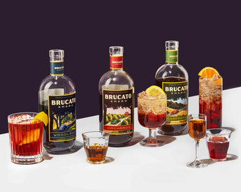 Brucato Amaro bottles and cocktails neatly styled on table in front of purple background